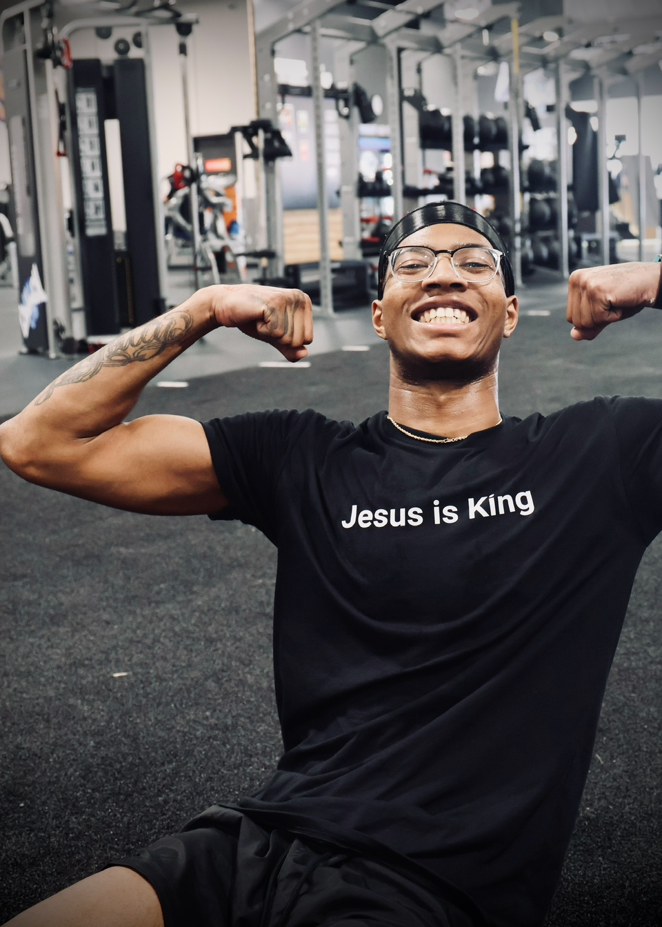 Person flexing muscles in a gym wearing a black t-shirt with 'Jesus is King' text.